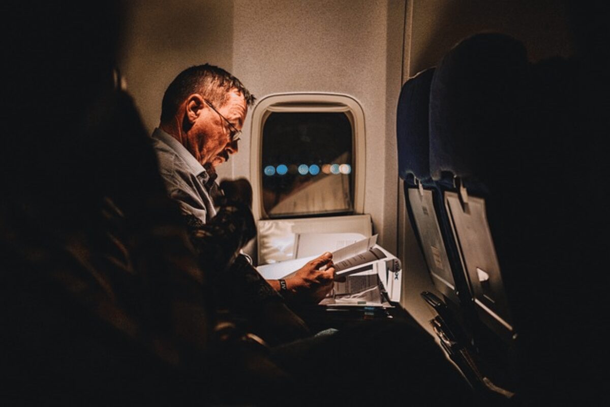 A man reading a book on a dimly lit airplane, with a focus on his illuminated face by the reading light.