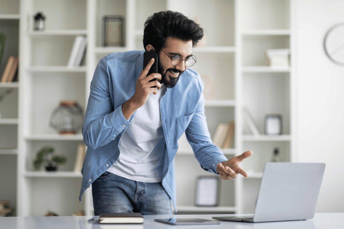 A man smiling and talking on the phone while gesturing with his hand at a laptop negotiating Air Miles points.